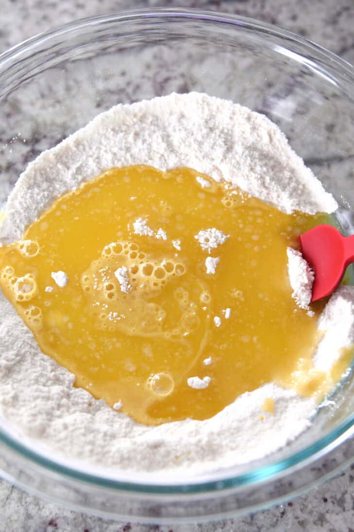 Top down view of glass bowl with flour, a lemon and egg mixture in the middle of the flour, and a red spatula on the side.
