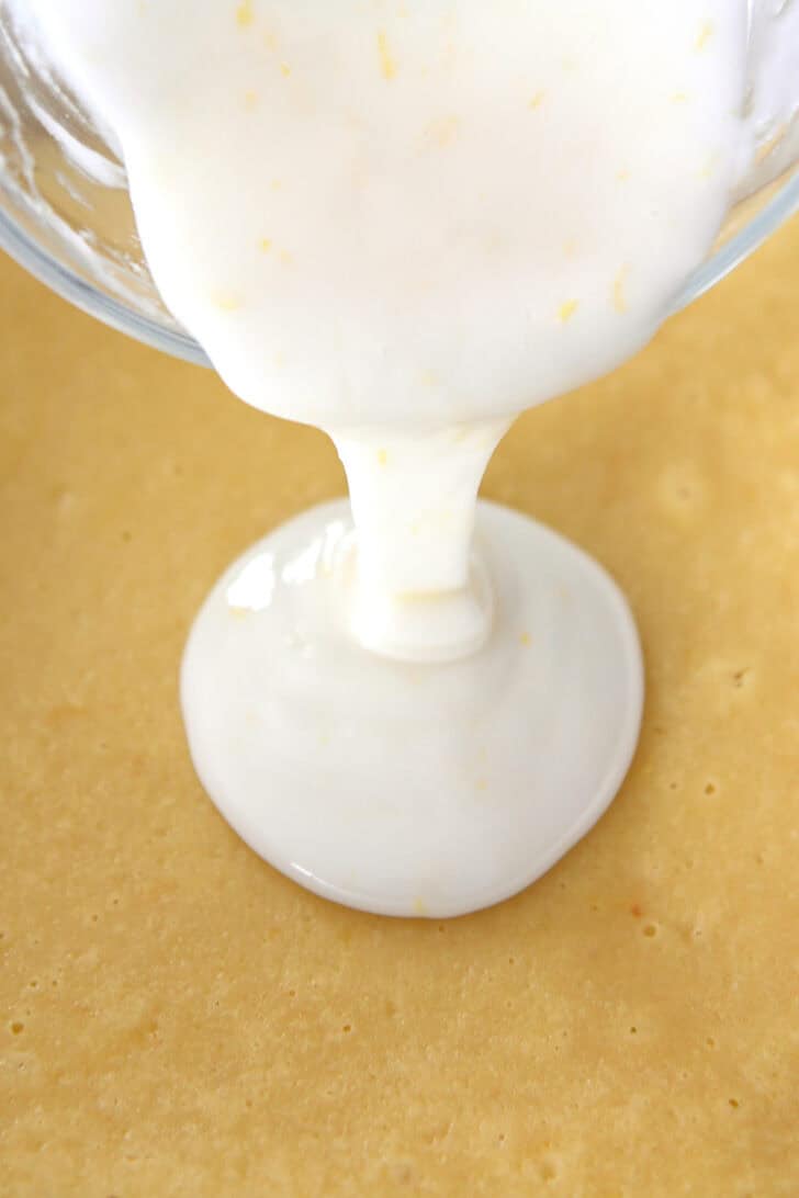 A glass bowl pouring a thick white glaze over a pan of lemon bars.