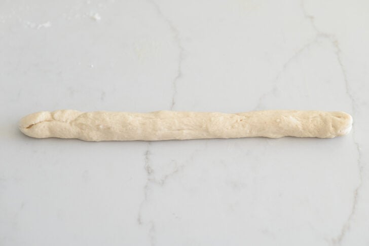 Dough formed into long, thin breadstick on white counter.