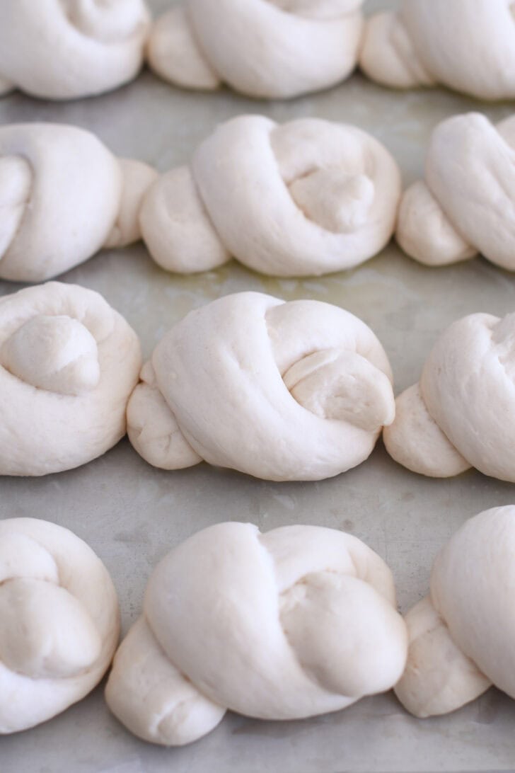 Nine risen breadstick dough knots on buttered baking sheet.
