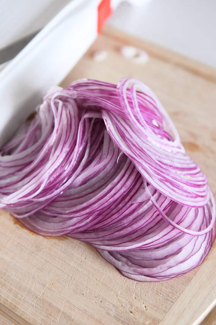 Thinly sliced red onions off a mandoline slicer on wood cutting board.