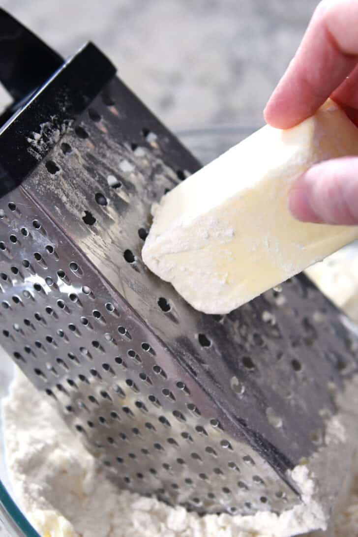 A hand grating a frozen cube of butter into a flour mixture in a glass bowl.