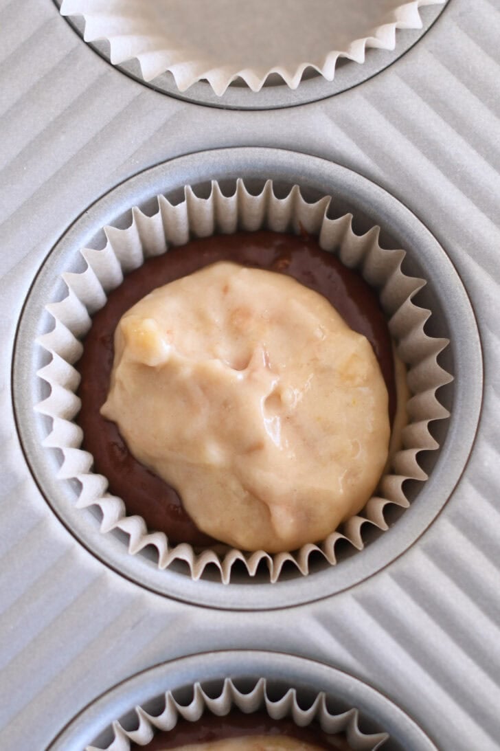 Muffin tin liner with scoop of banana muffin batter on top of chocolate banana muffin batter.
