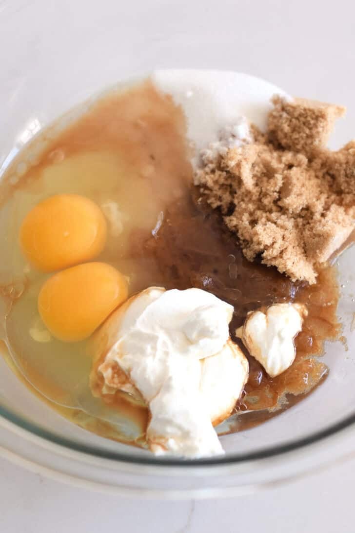 Glass bowl with eggs, sour cream, brown sugar, oil and granulated sugar.