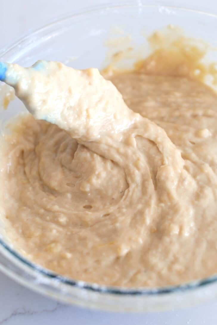 Spatula scooping and swirling banana muffin batter in glass bowl.