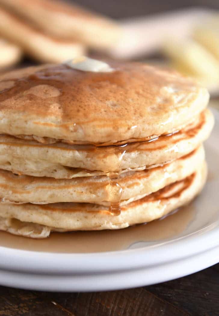 Stack of four pancakes on white plates with syrup drizzled down the side.