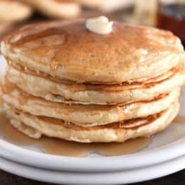 Stack of four pancakes on white plates with butter and syrup.