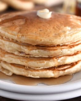 Stack of four pancakes on white plates with butter and syrup.