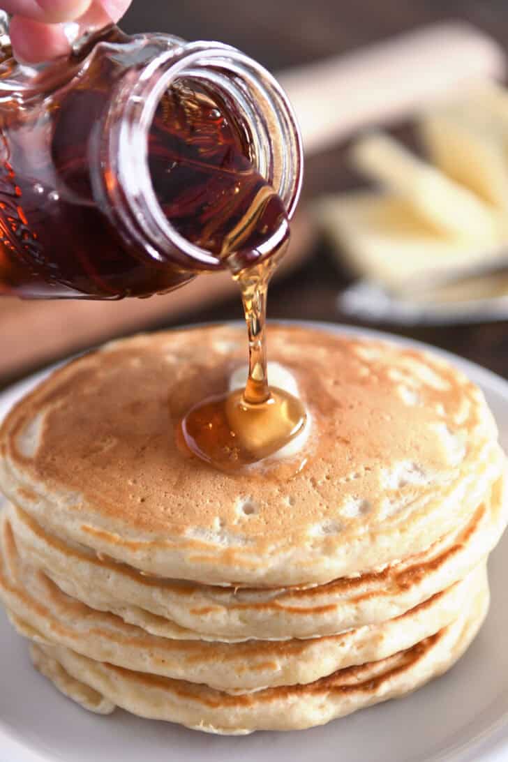 Pouring syrup from glass jar onto stack of four pancakes on white plates.
