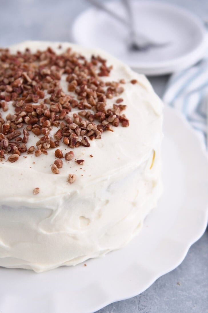 Side view of frosted layer carrot cake with toasted pecans on white platter.