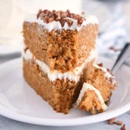 Slice of carrot layer cake on white plate with bite taken out on fork.