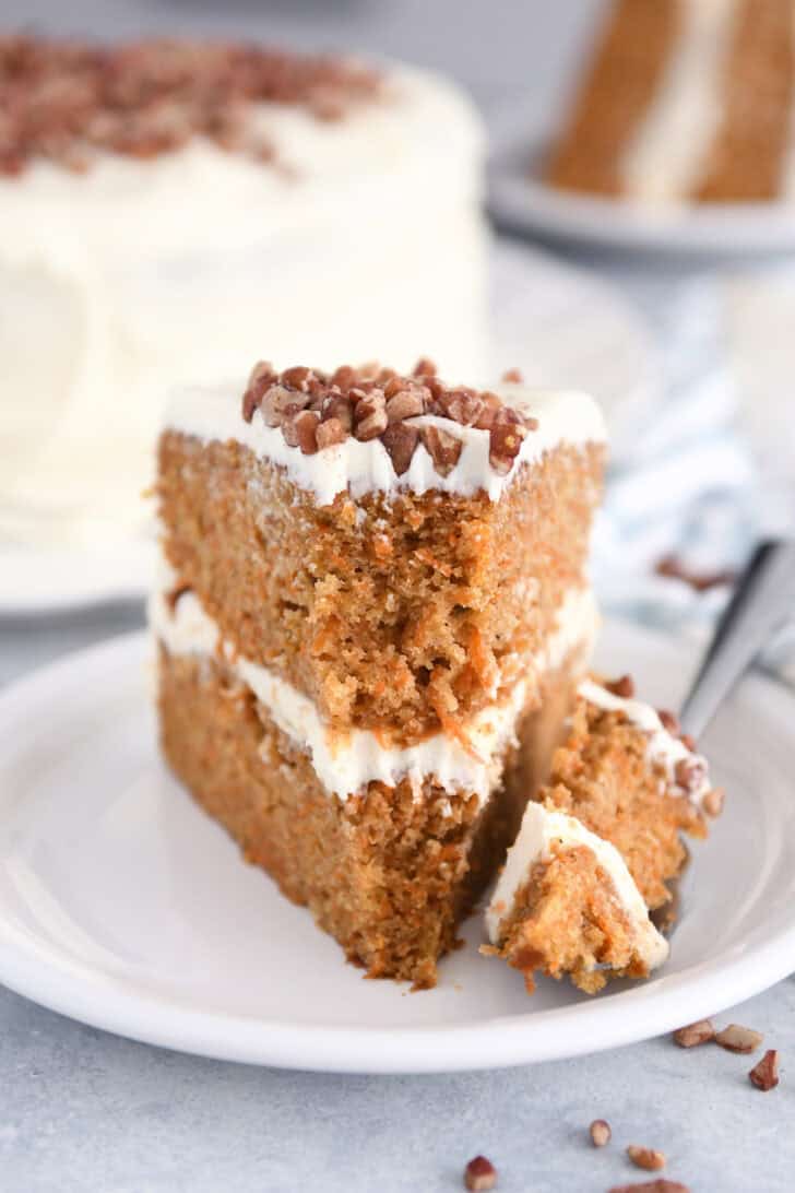 Slice of carrot cake on white plate with bite taken out on fork next to the slice of cake.