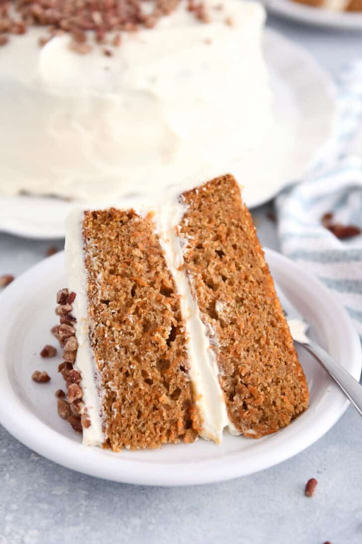 Slice of carrot cake on white plate with frosting and pecans.
