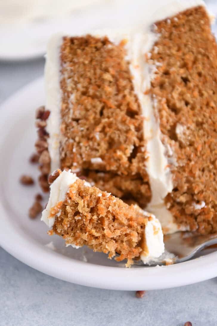 Bite of carrot cake on fork with piece of carrot cake on white plate.