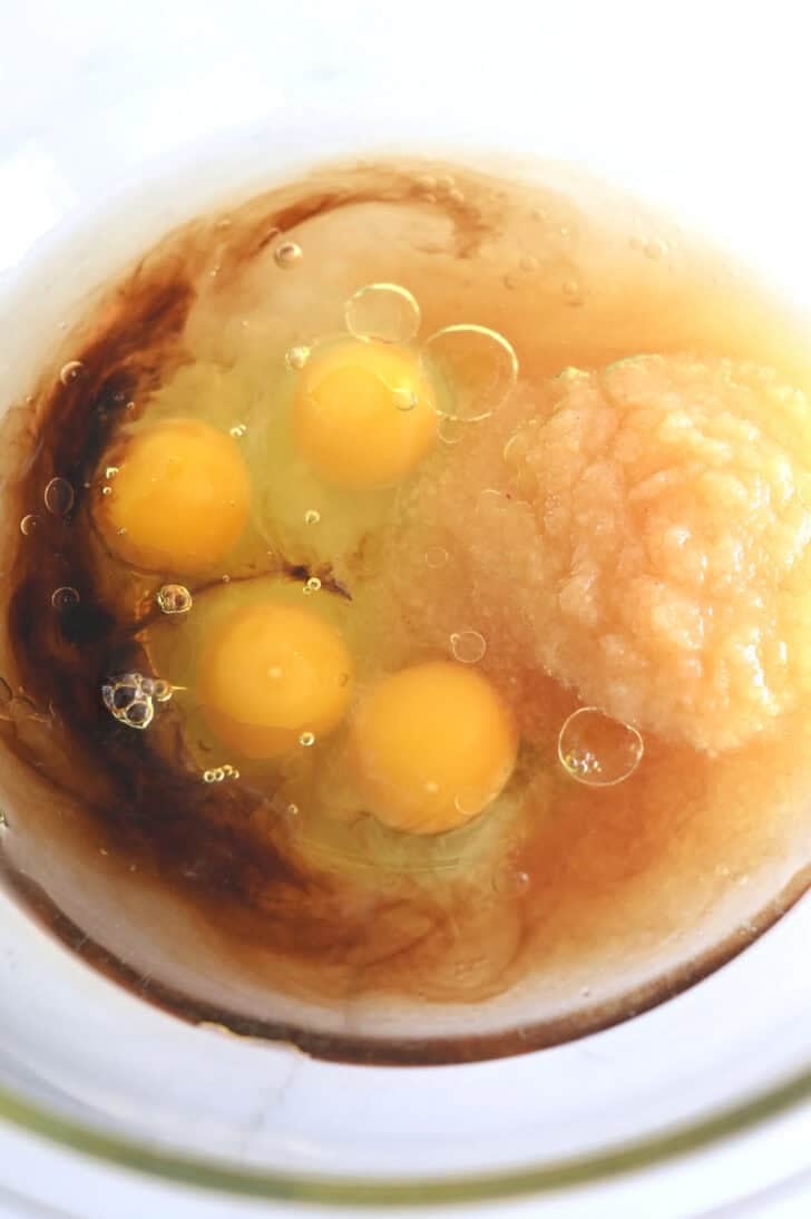 Top down view of a glass bowl with raw eggs, vanilla, applesauce, and oil.