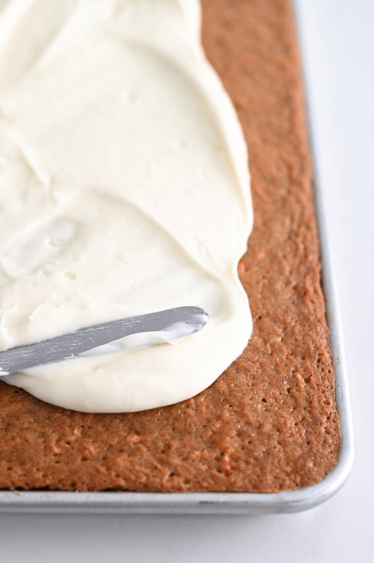 A sheet pan of carrot cake getting frosted with cream cheese frosting.