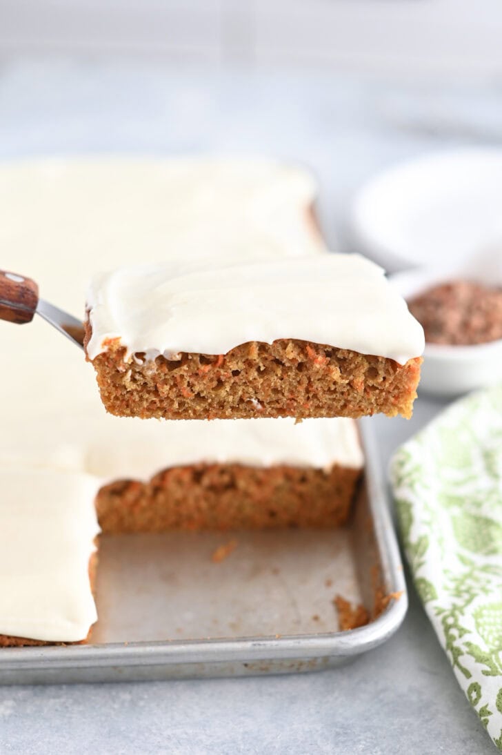 A metal spatula holding a piece of frosted carrot sheet cake above a sheet pan of cake.