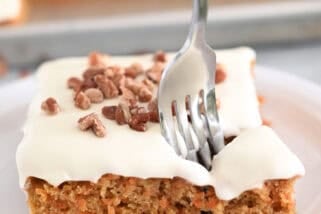 A piece of cream cheese frosted carrot sheet cake sprinkled with pecans on a white plate. A fork is taking a bite out of the cake.