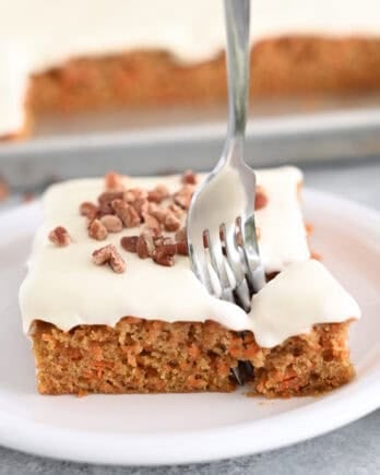 A piece of cream cheese frosted carrot sheet cake sprinkled with pecans on a white plate. A fork is taking a bite out of the cake.