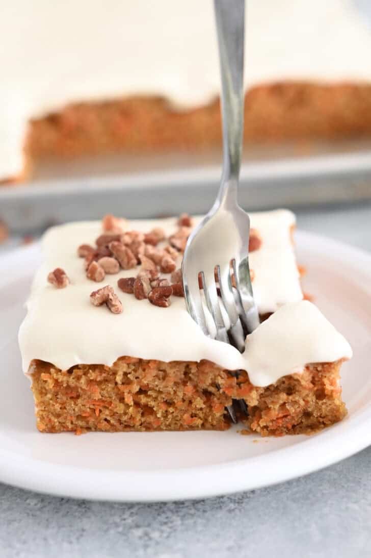 A piece of cream cheese frosted carrot sheet cake sprinkled with pecans on a white plate. A fork is taking a bite out of the cake.