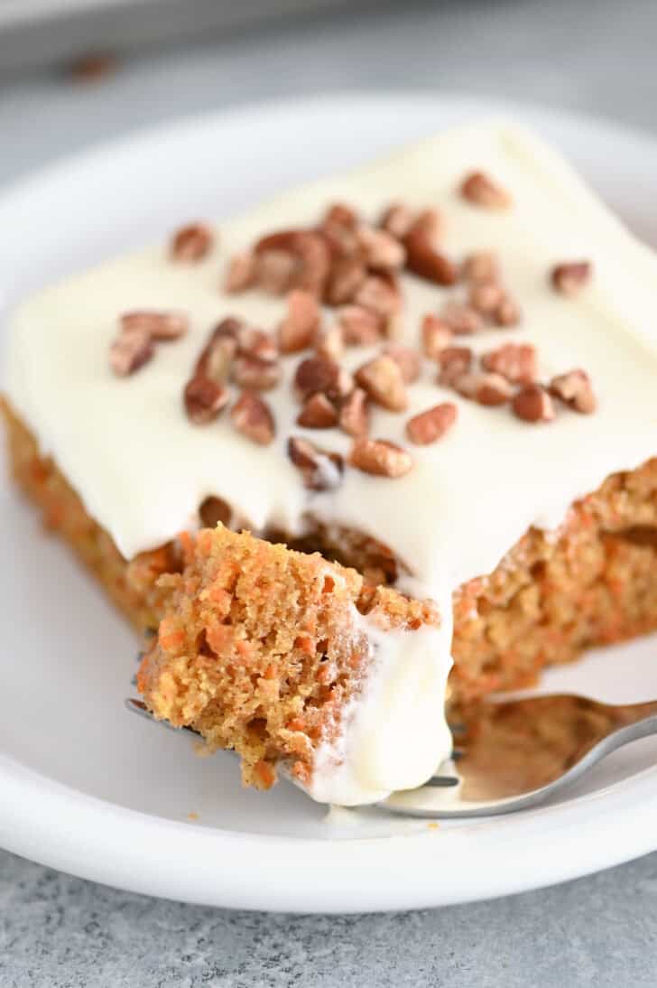 A piece of cream cheese frosted carrot sheet cake sprinkled with pecans on a white plate. A fork is taking a bite out of the cake.
