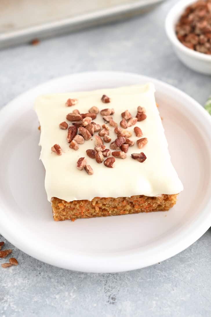 Top down view of a white plate with a piece of pecan sprinkled carrot sheet cake with white cream cheese frosting.