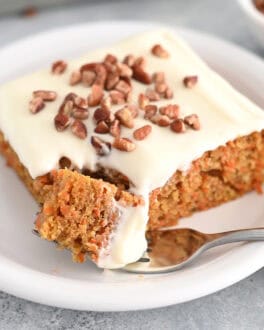 A piece of cream cheese frosted carrot sheet cake sprinkled with pecans on a white plate. A fork is taking a bite out of the cake.
