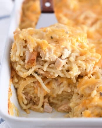 Spatula holding square of cheesy chicken spaghetti in white pan with the rest of the casserole.
