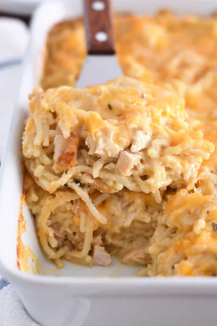 Spatula holding square of cheesy chicken spaghetti in white pan with the rest of the casserole.