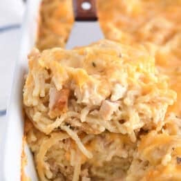 Spatula holding square of cheesy chicken spaghetti in white pan with the rest of the casserole.