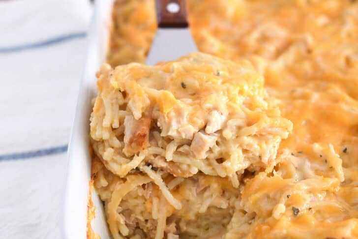 Spatula holding square of cheesy chicken spaghetti in white pan with the rest of the casserole.