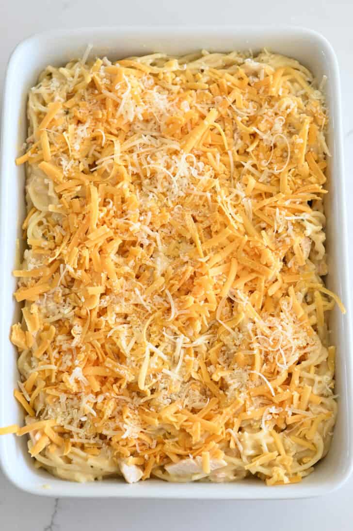 Top down view of shredded cheddar and Parmesan cheeses on top of cooked spaghetti with cheese sauce and cooked chicken.