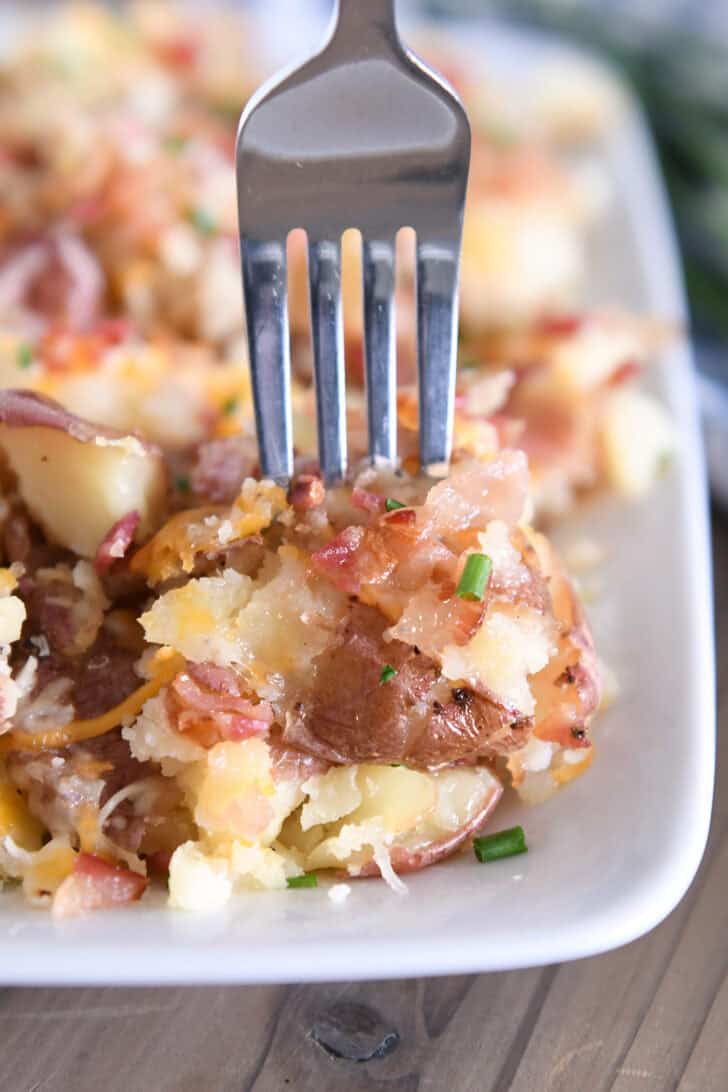 Fork piercing bite of cheesy smashed potatoes on white tray.
