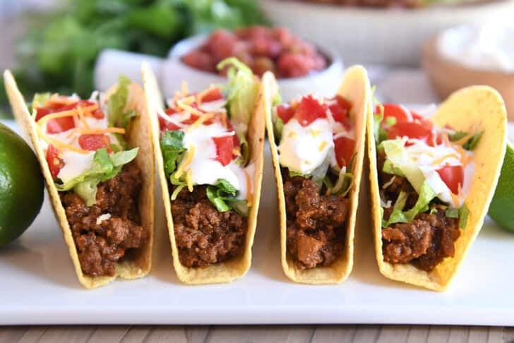 Four crispy-shelled ground beef tacos filled with toppings on a white platter.