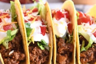 Four crispy-shelled ground beef tacos filled with toppings on a white platter.