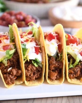Four crispy-shelled ground beef tacos filled with toppings on a white platter.