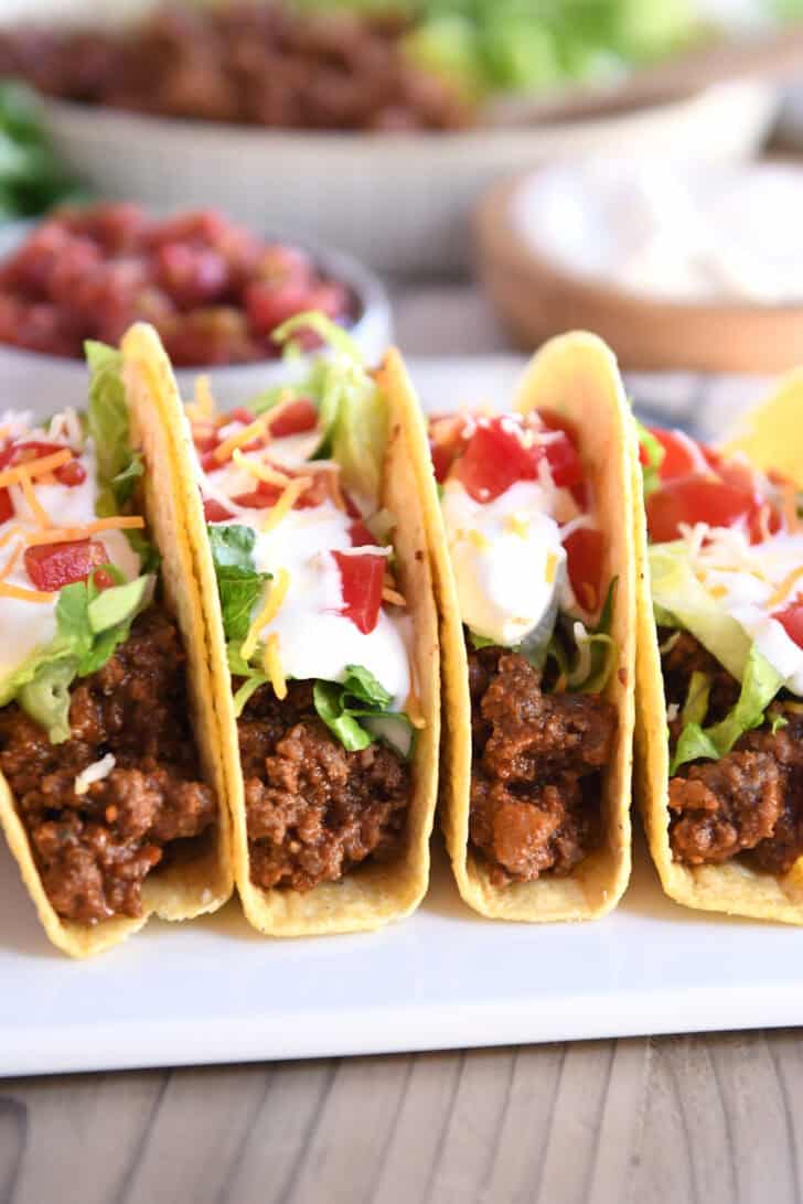 Four crispy-shelled ground beef tacos filled with toppings on a white platter.