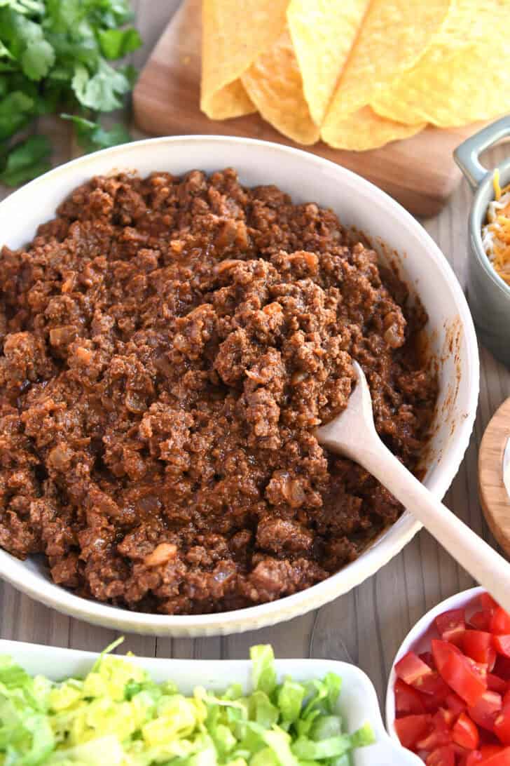 Top down view of a white bowl filled with seasoned ground taco meat. Taco toppings are in bowls around the meat.