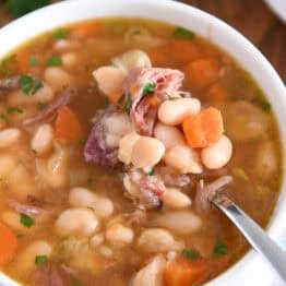 White bowl with handles with spoon lifting bite of white beans, carrots and ham.