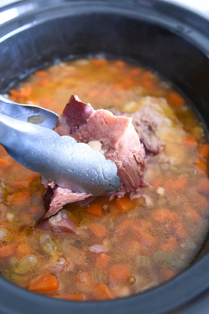 Lifting ham bones with tongs out of slow cooker with white beans, broth and veggies.