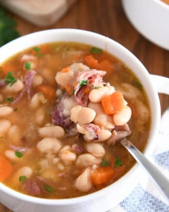Top down view of ham and bean soup in white bowl with handles with spoon lifting bite of white beans, carrots and ham.
