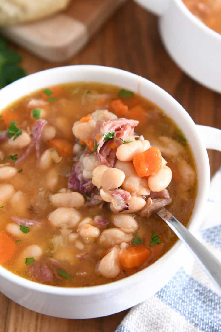 Top down view of ham and bean soup in white bowl with handles with spoon lifting bite of white beans, carrots and ham.