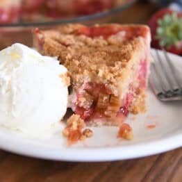 Half eaten slice of strawberry rhubarb pie on white plate with vanilla ice cream.