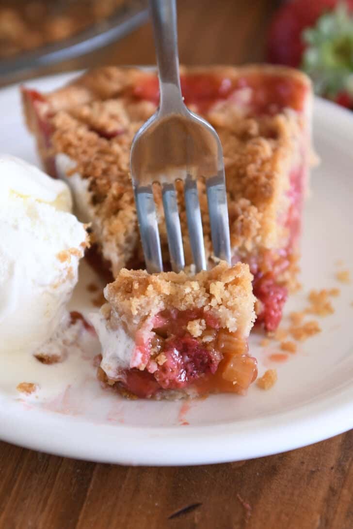 Fork with bite of strawberry rhubarb pie on white plate with vanilla ice cream.