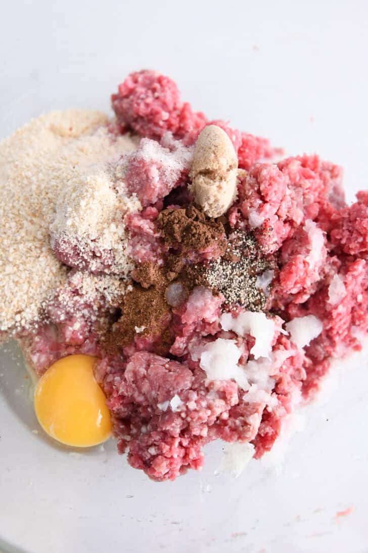 Glass bowl with ground beef, grated onion, egg, pepper, nutmeg, brown sugar and bread crumbs.