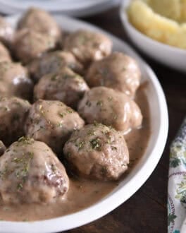 White platter with lots of cooked meatballs in brown gravy and dried parsley on top.