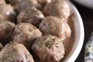 White platter with lots of cooked meatballs in brown gravy and dried parsley on top.