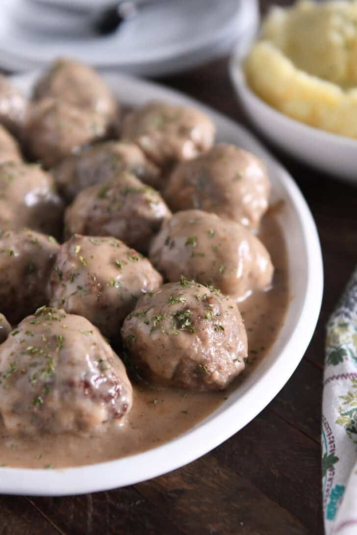 White platter with lots of cooked meatballs in brown gravy and dried parsley on top.