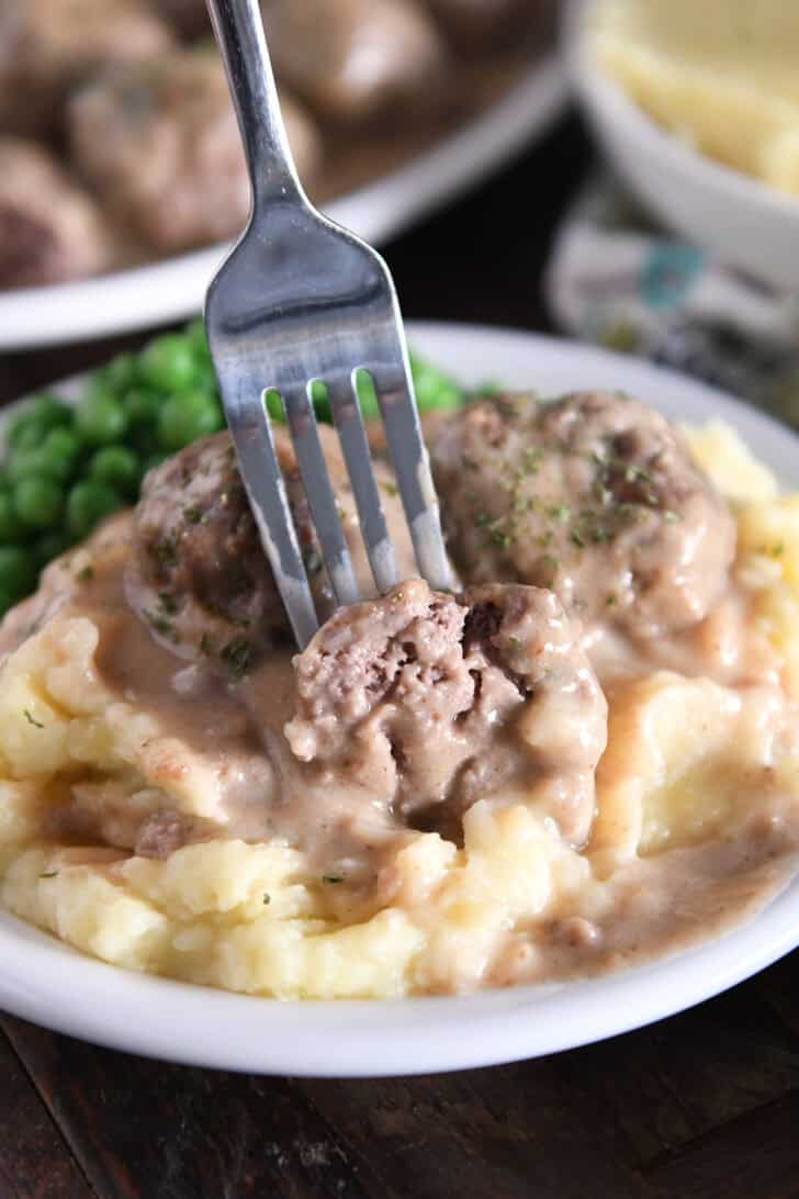Fork piercing half meatball on top of creamy mashed potatoes smothered in brown gravy.