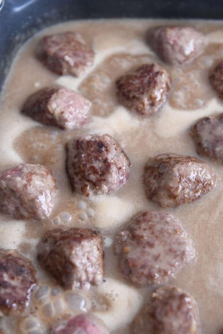 Browned meatballs cooking in brown gravy sauce in electric skillet.